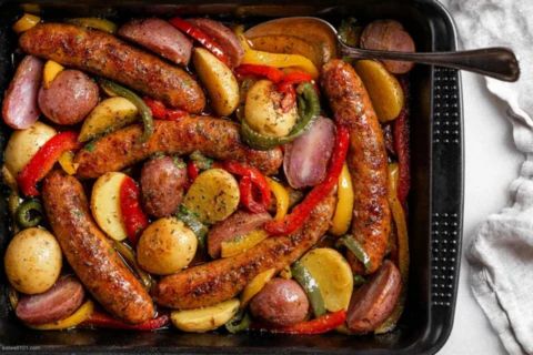 Oven-baked Italian sausages served with roasted peppers, onions, and potatoes, alongside pasta, sandwiches, and fresh salad, showcasing versatile meal options.
